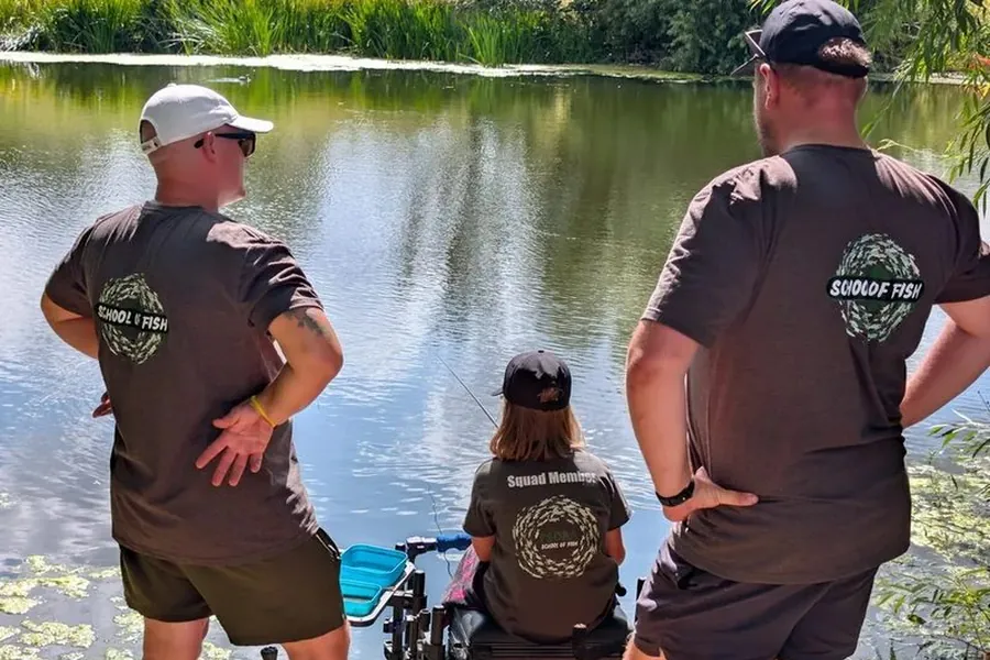 Two School Of Fish coaches standing behind a junior squad member as she fishes a lake